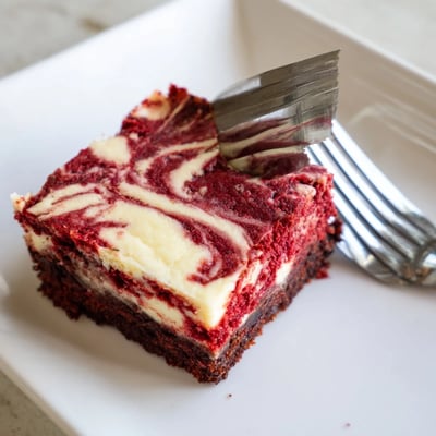 A slice of Red Velvet Cheesecake Brownies reveals a fudgy red velvet base and creamy cheesecake layers on a white plate.